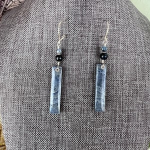 May include: A pair of dangle earrings with silver-tone hooks. Each earring features a rectangular, speckled, gray-blue stone pendant. Above the pendant are black and gray beads.