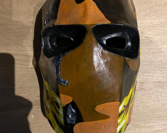 May include: A handmade mask with a camouflage design. The mask features a combination of black, orange, and yellow paint. The eye openings are black. The mask is designed to cover the entire face.