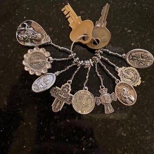 May include: A silver-toned metal keyring featuring two standard brass keys and a collection of eight religious charms. The charms include various circular and cross-shaped pendants depicting figures and symbols, all attached to a central metal ring.