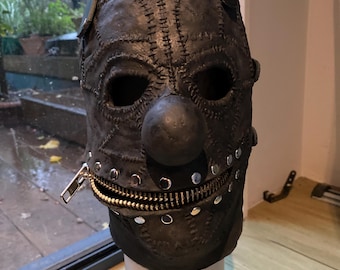 May include: A dark gray full-head mask featuring a prominent round nose, two eye openings, and a horizontal mouth area secured with a metal zipper. The surface is textured with visible stitched seams and accented with silver-toned metal studs along the jawline.