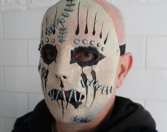 May include: A white Halloween mask with black stitching and silver accents. The mask has a stitched mouth and eyes, and a textured surface.