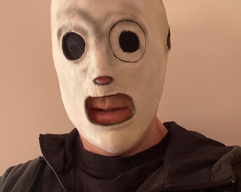 May include: A full-head mask featuring a smooth, off-white surface with a distinct circular black outline around the left eye, dark mesh eye coverings, a small red nose detail, and a wide open mouth cutout, worn over a black zip-up hooded jacket.