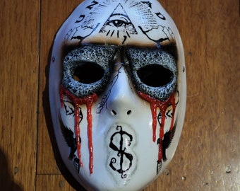 May include: White plastic mask with a black and red design. The mask has a black eye hole design, a black dollar sign, and the words "UNDEAD" written on the top. The mask has red paint dripping from the eye holes.