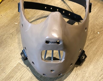 May include: A tan-colored mask with a black leather strap and metal bars over the mouth. The mask has multiple small holes and is designed to fit over the face. The mask is a prop.