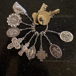 May include: A silver-toned metal keyring featuring two brass keys and a collection of nine religious charms. The charms include various oval and cross-shaped pendants depicting figures, symbols, and the text "Our Lady of Perpetual Help".