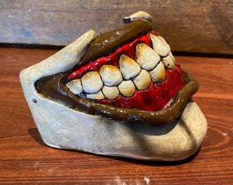 May include: A ceramic sculpture of a wide open mouth with large, off-white teeth, brown gums, and a red interior. The sculpture is set on a wooden surface.