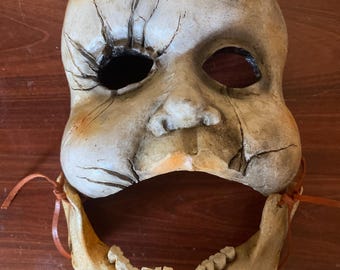 May include: A distressed, off-white mask with a cracked surface and a missing eye. The mask features a sculpted face with a skeletal jaw attached with leather ties. The jaw has visible teeth, creating a haunting and eerie appearance.