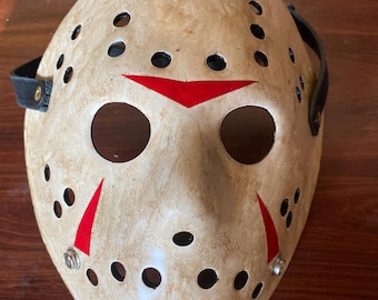 May include: A cream-colored hockey mask with black eye holes and numerous small, evenly spaced holes. Red triangles are painted on the forehead and cheeks. The mask has black straps and is likely for costume or display.