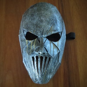 May include: A distressed, metallic-looking mask with a menacing design. The mask features large eye openings, a prominent nose, and a mouth area with vertical slits. It has a weathered appearance with streaks of gold and a black strap.