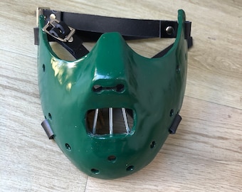 May include: A green leather mask with a metal cage over the mouth and a black leather strap. The mask has a series of small holes around the edges.