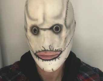May include: A full-head mask featuring a pale, off-white complexion with dark shading around the eye sockets. The mask displays prominent black stitched detailing along the sides and mouth area, with two dark, circular eye openings and pink lips.