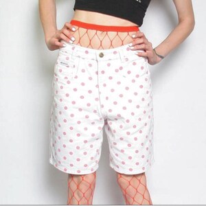 May include: White denim shorts with a pink polka dot pattern. The shorts have a button closure and pockets. The model is wearing red fishnet stockings and a black top.