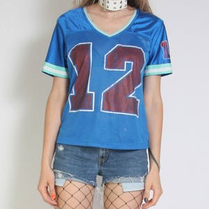 May include: A blue jersey with the number 12 printed on the front. The jersey has white and green trim on the sleeves and collar. The model is wearing the jersey with denim shorts and fishnet stockings.