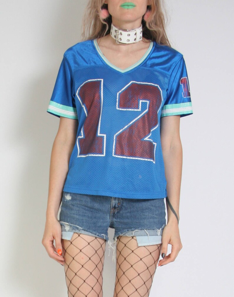 May include: A blue jersey with the number 12 printed on the front. The jersey has white and green trim on the sleeves and collar. The model is wearing the jersey with denim shorts and fishnet stockings.