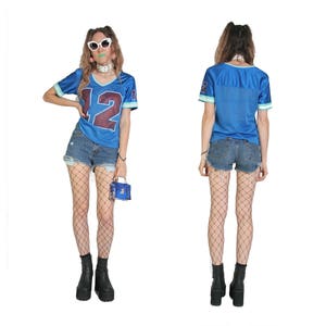May include: A blue jersey with the number 12 on the front, worn with denim shorts and fishnet tights. The person is wearing white sunglasses and a silver necklace.