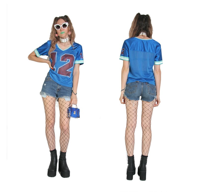 May include: A blue jersey with the number 12 on the front, worn with denim shorts and fishnet tights. The person is wearing white sunglasses and a silver necklace.