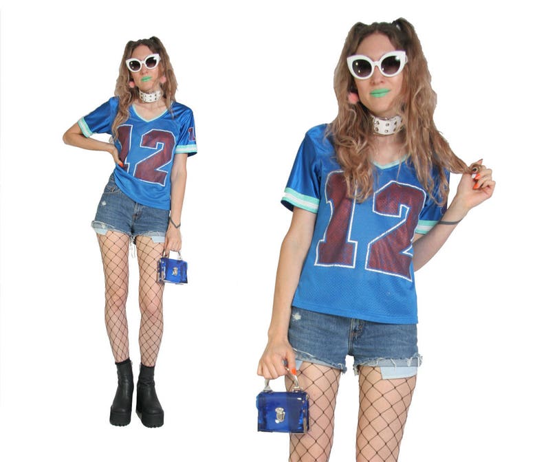 May include: A woman wearing a blue jersey with the number 12 on it, white sunglasses, and fishnet stockings. She is holding a small blue box purse.