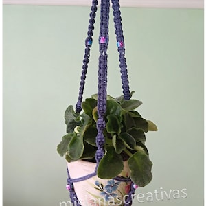 Puede incluir: Un macetero de macramé morado con una pequeña planta en maceta. El macetero tiene cuentas en tonos rosa y azul. La maceta tiene un diseño floral y el texto "mislanascreativas etsy.com".