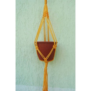 May include: A yellow macrame plant hanger with a brown terracotta pot hanging from it. The hanger has a long tassel at the bottom.