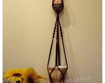 Pode incluir: Um suporte de macramé castanho para plantas com dois vasos. O vaso superior é um vaso pequeno, redondo, laranja e preto. O vaso inferior é um vaso maior, redondo, castanho e branco. O suporte tem uma franja comprida na parte inferior.