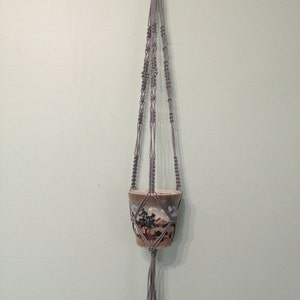 May include: A gray macrame plant hanger with a pot featuring a landscape scene. The hanger is made of cotton cord and has a diamond pattern.