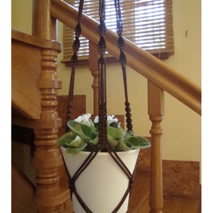 May include: A brown macrame plant hanger with a white pot and green plant hanging from a wooden staircase railing.