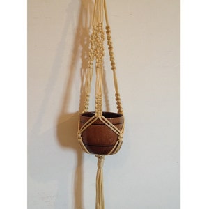 Pode incluir: Um suporte de plantas de macramé com um vaso castanho pendurado nele. O suporte é feito de corda de cor creme e tem uma borla comprida.