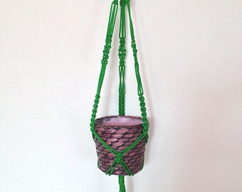 Pode incluir: Um suporte de plantas de macramê verde com um vaso de vime marrom pendurado.