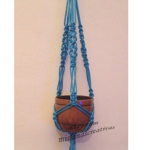Può includere: Un portavasi in macramè turchese con un vaso in legno. Il portavasi è realizzato con una tecnica di annodatura e ha una frangia in basso. Il testo "etsy.com mislanascreativas" è visibile nell'immagine.