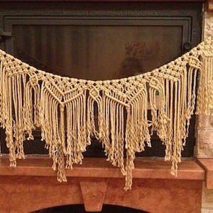 May include: A beige macrame garland with a geometric pattern hangs above a fireplace. The garland has long fringe and is made of natural fibres.