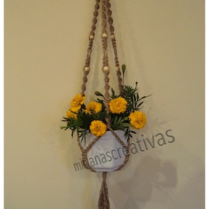May include: A macrame plant hanger with wooden beads, holding a white pot with yellow flowers. The hanger has a long tassel at the bottom. The text "misanascreativas" is visible in the lower right corner of the image.
