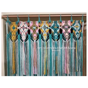 Könnte beinhalten: Ein Wandbehang aus Makramee mit einem geometrischen Muster in Türkis, Gelb, Rosa und Grau. Der Behang hat unten eine Fransenborte mit Quasten.