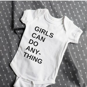 Pu&ograve; includere: Body bianco per neonato con testo nero che recita "Girls Can Do Anything".
