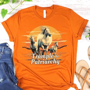 May include: Orange t-shirt featuring a graphic of three white horses running against a sunset backdrop. The text "Trample the Patriarchy" is below the horses. The shirt is styled with blue jeans and sandals.
