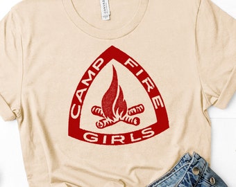 May include: A beige t-shirt with a red triangle logo that says "Camp Fire Girls" with a stylized image of a campfire inside the triangle.