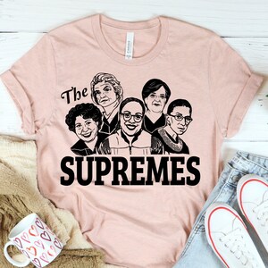 Pu&ograve; includere: Una maglietta rosa con una grafica nera di cinque volti di donne e il testo "The Supremes".