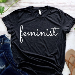 May include: A dark gray t-shirt with the word "feminist" written in white cursive script.