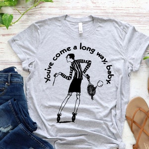 May include: Gray t-shirt with a black and white illustration of a woman holding a tennis racket. The text "You've come a long way, baby" is printed on the shirt.