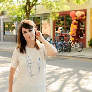 May include: A woman wearing a white t-shirt with a blue illustration of two people. The illustration shows one person pointing at the other. The woman is standing on a street in front of a store with a sign that says "LIT CO. KICKS".