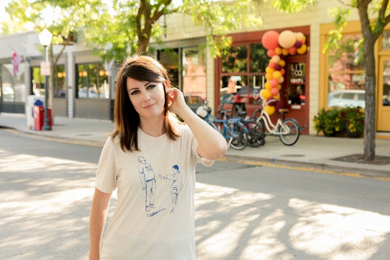 May include: A woman wearing a white t-shirt with a blue illustration of two people. The illustration shows one person pointing at the other. The woman is standing on a street in front of a store with a sign that says "LIT CO. KICKS".