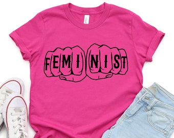 May include: Pink t-shirt with a black graphic of two fists with the word "FEMINIST" spelled out on the knuckles.