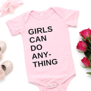 Pu&ograve; includere: Body rosa per neonato con il testo "GIRLS CAN DO ANY-THING" in lettere nere. Il body &egrave; circondato da accessori rosa, tra cui una fascia per capelli e scarpe.