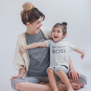 May include: A woman and a young girl are sitting together. The woman is wearing a gray t-shirt with a list of women's names printed on it. The girl is wearing a white t-shirt with the words "Bossy Boss" printed on it. They are both smiling and looking at each other.