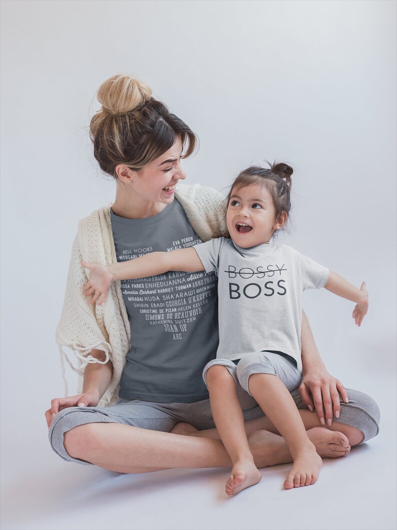 May include: A woman and a young girl are sitting together. The woman is wearing a gray t-shirt with a list of women's names printed on it. The girl is wearing a white t-shirt with the words "Bossy Boss" printed on it. They are both smiling and looking at each other.