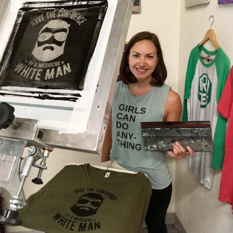 K&ouml;nnte beinhalten: Eine Frau tr&auml;gt ein hellblaues Tanktop mit dem Aufdruck "Girls Can Do Anything". Sie h&auml;lt einen Siebdruckrahmen mit einem schwarz-wei&szlig;en Design eines Mannes mit Bart und Sonnenbrille. Das Design lautet "Have the Confidence of a Mediocre White Man".