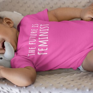 Pu&ograve; includere: Un body rosa per neonato con il testo "THE FUTURE IS FEMINIST" in bianco.