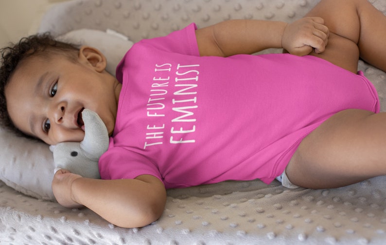 Pu&ograve; includere: Un body rosa per neonato con il testo "THE FUTURE IS FEMINIST" in bianco.