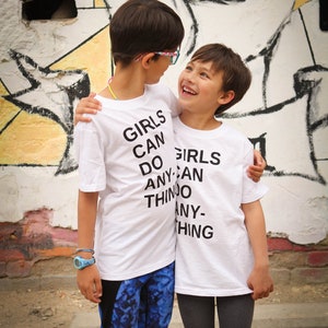 K&ouml;nnte beinhalten: Zwei Kinder tragen wei&szlig;e T-Shirts mit dem Aufdruck "Girls Can Do Anything". Das Kind links tr&auml;gt blaue Leggings, das Kind rechts graue Leggings. Sie stehen vor einer Graffiti-Wand.