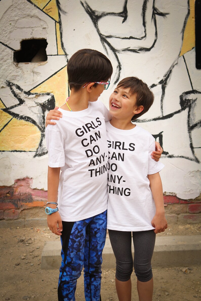 K&ouml;nnte beinhalten: Zwei Kinder tragen wei&szlig;e T-Shirts mit dem Aufdruck "Girls Can Do Anything". Das Kind links tr&auml;gt blaue Leggings, das Kind rechts graue Leggings. Sie stehen vor einer Graffiti-Wand.