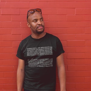 Puede incluir: Una persona lleva una camiseta negra con texto blanco que dice "Los derechos de las mujeres son los derechos de los hombres, los derechos trans son los derechos de las personas con discapacidad, los derechos de los homosexuales son los derechos de los trabajadores, los derechos religiosos son los derechos civiles, los derechos de los refugiados son los derechos de las mujeres, los derechos de los hombres son los derechos trans". La persona est&aacute; de pie frente a una pared de ladrillo rojo.